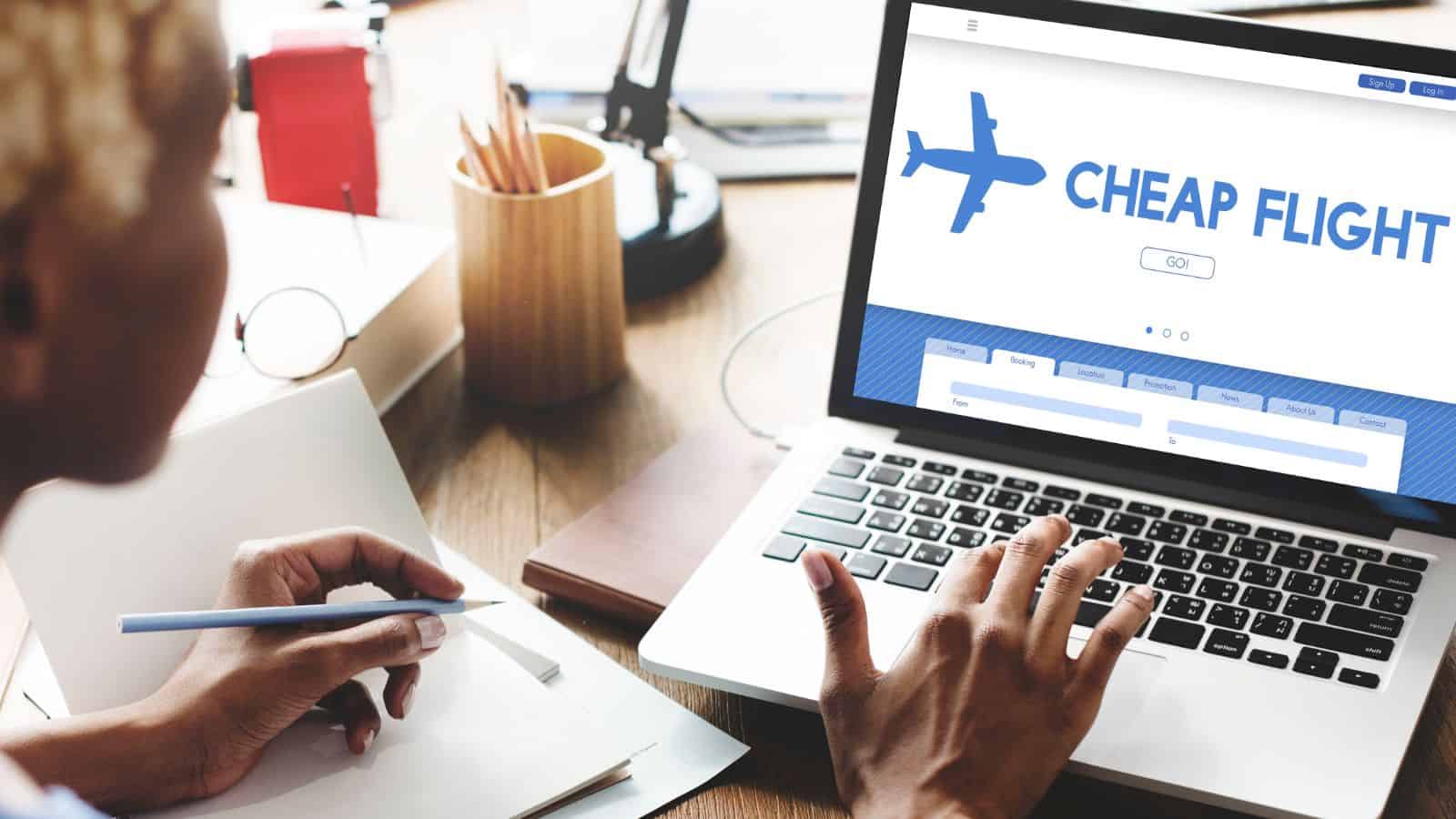 A person is using a laptop displaying a website with the words "CHEAP FLIGHT" and a search button. They are sitting at a wooden desk with a notebook, pen, plant, and various office supplies.