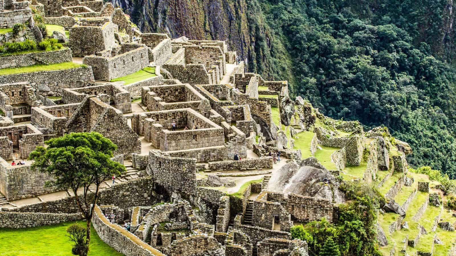 Ruins of Machu Picchu, an ancient Incan city, featuring stone structures and terraces on a mountainside. Lush greenery surrounds the site, with a dense forest visible in the background. Some stairways and paths are evident among the stonework.
