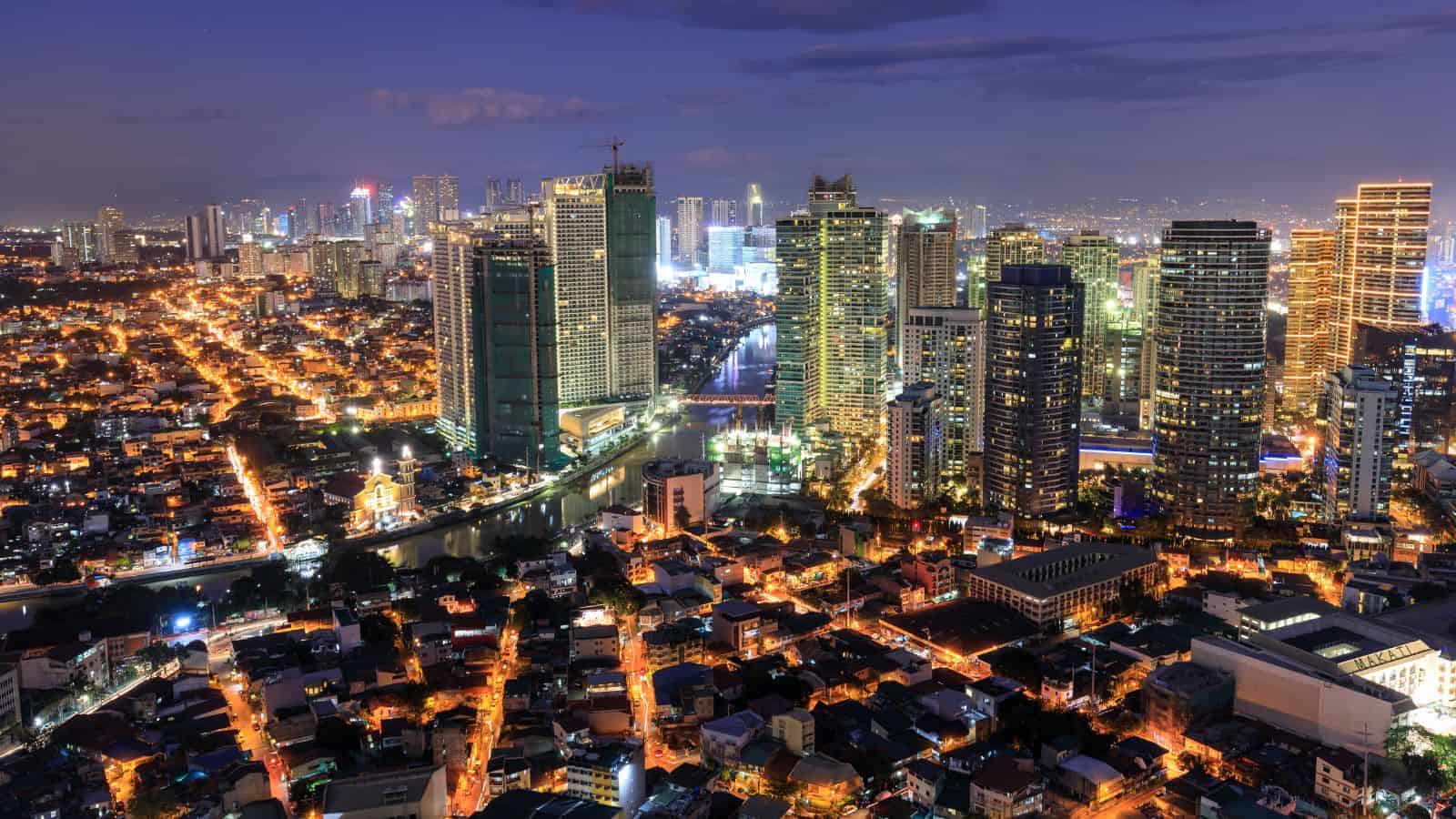Aerial view of a cityscape at night, featuring a mix of tall skyscrapers and smaller buildings. Streets are illuminated with orange lights, and the skyline is bustling with bright lights, set against a dark blue sky.