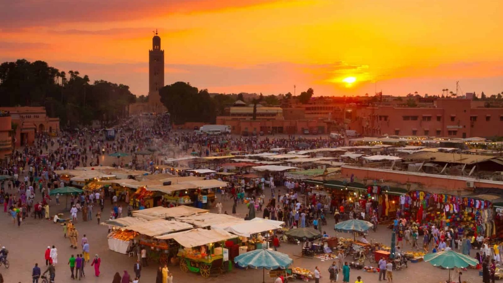 Sunset in Marrakech shows crowds at a vibrant outdoor market with colorful stalls and a mosque tower in the background.