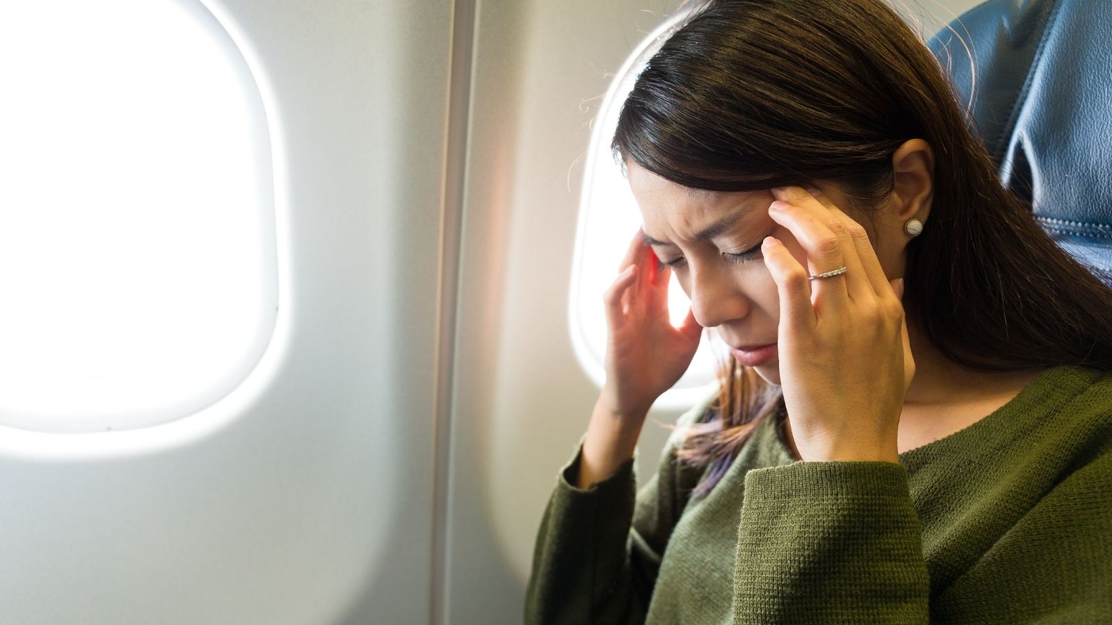 A woman on an airplane holds her head with both hands, appearing to have a headache or discomfort.