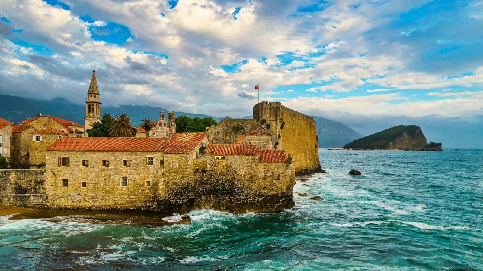Stone fortress and red-roofed old town by the sea, waves against walls, under a cloudy sky with a distant island.