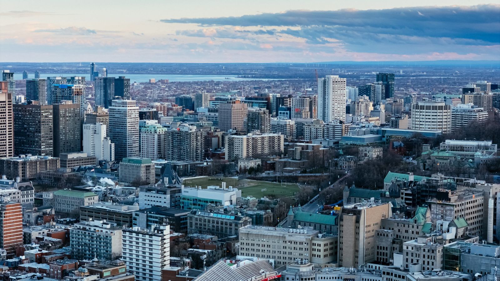 Aerial view of a city with tall buildings, mid-rises, and green spaces beneath a partly cloudy sky.