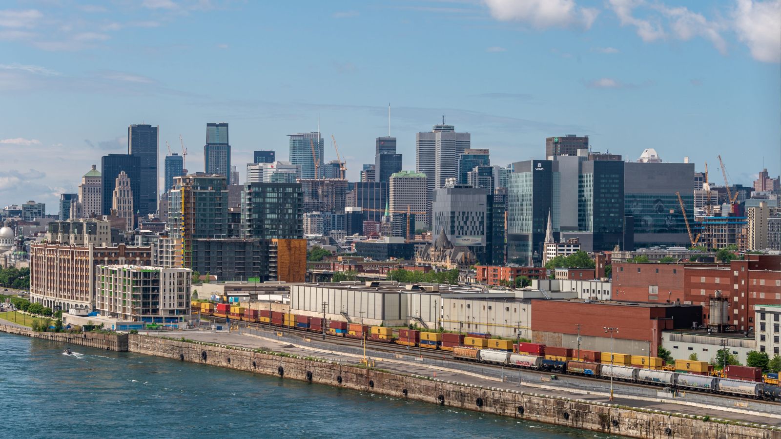 A city skyline of modern and historic buildings overlooks a waterfront, with a cargo train of colorful containers in front.