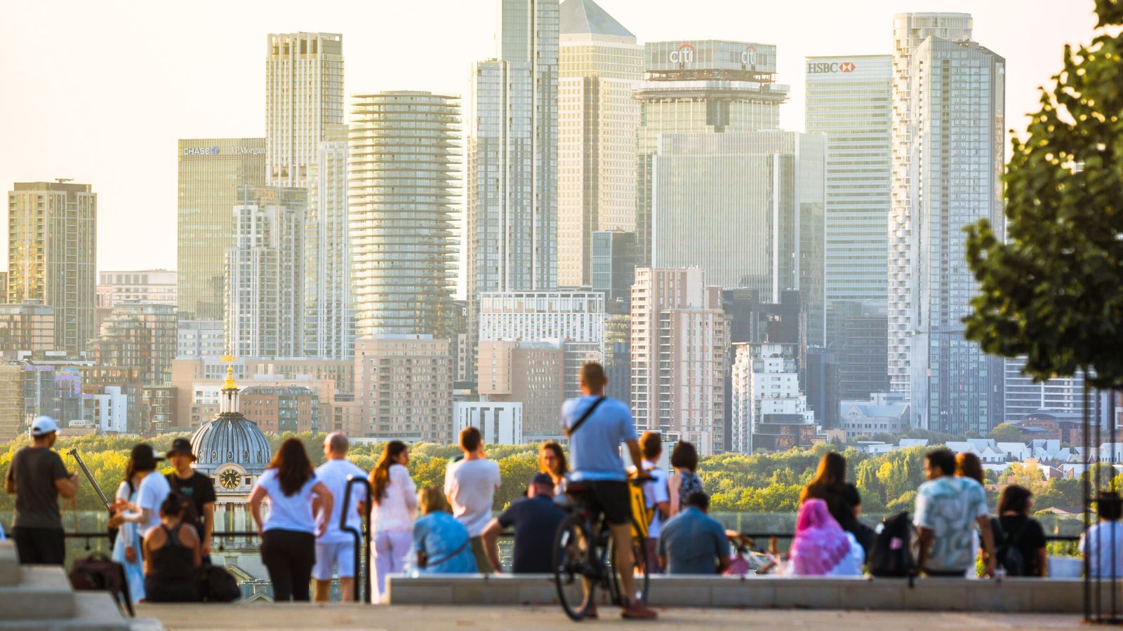 People gather outdoors with a modern city skyline of tall glass buildings and green trees visible in the background.