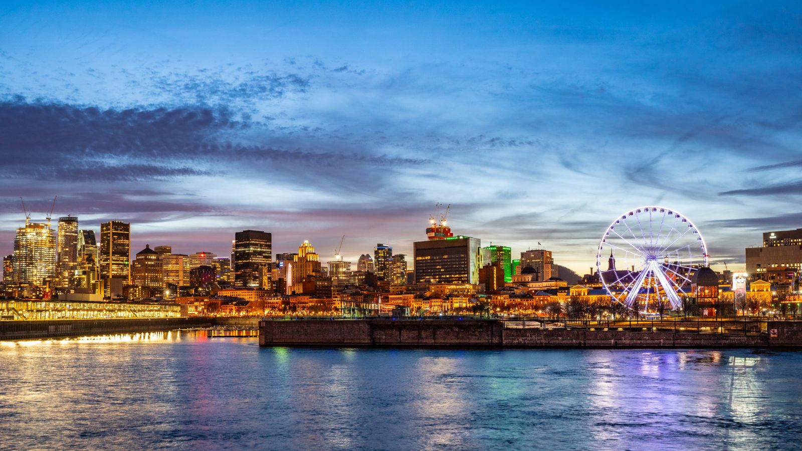 Dusk city skyline with lit buildings and a Ferris wheel reflected on water.