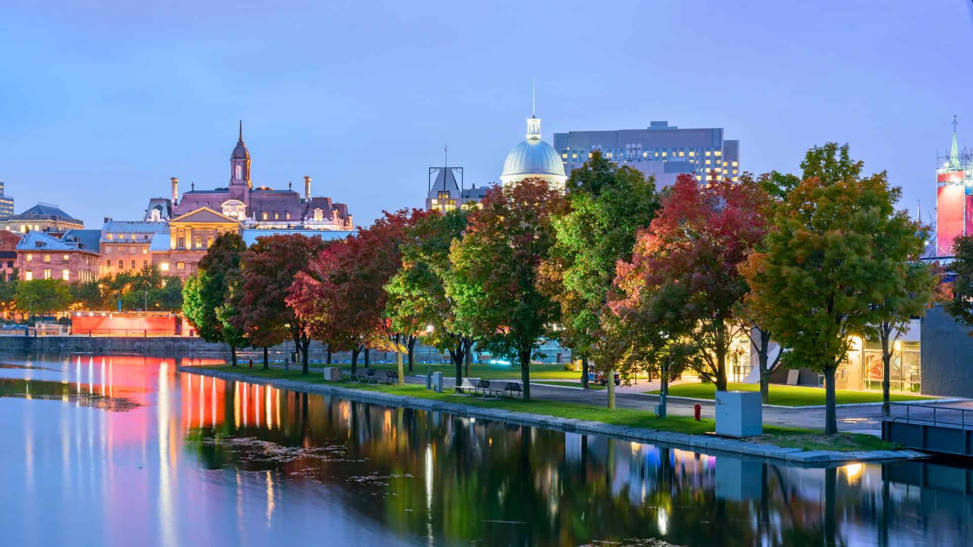 Colorful autumn trees line a city waterfront at dusk, with lit historic buildings reflected in the calm water.