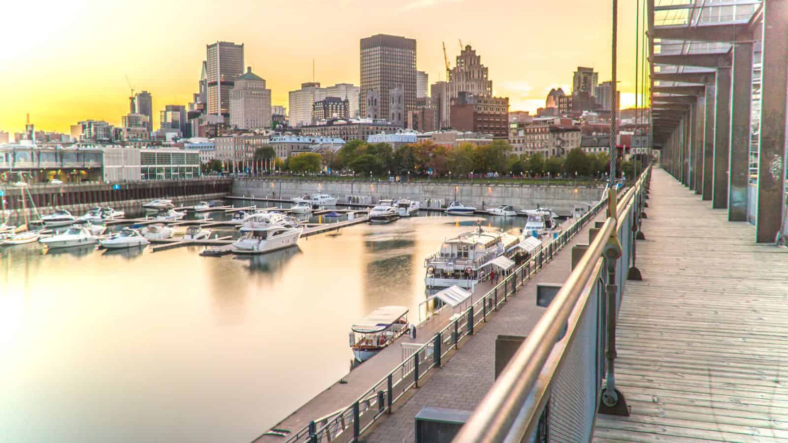 Docked boats at a marina beside a boardwalk, city skyline and tall buildings silhouetted against a sunset in the background.
