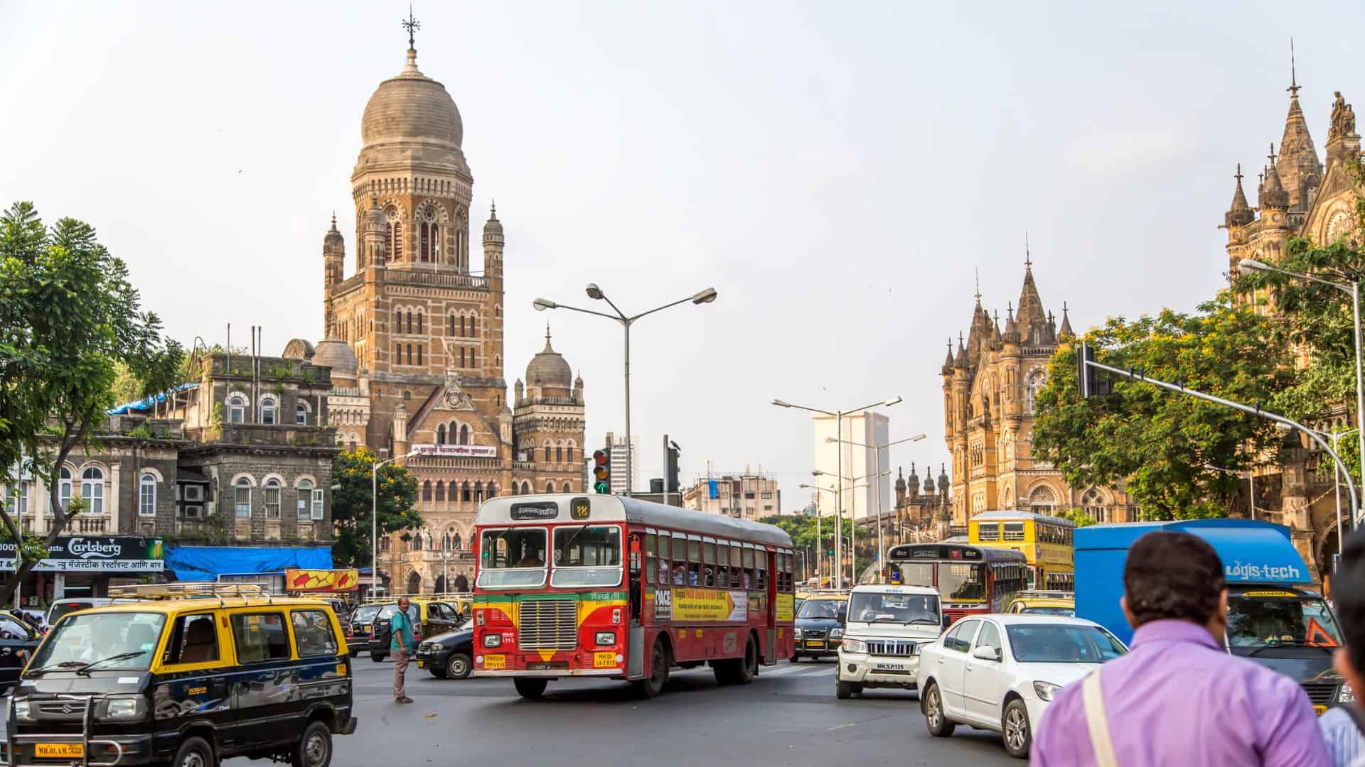 Busy Mumbai street with red bus, taxis, and historic colonial buildings in the background, daytime.