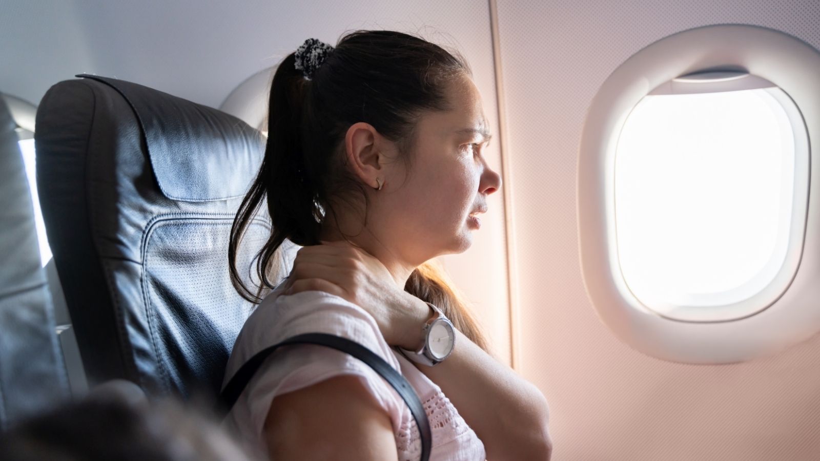 A concerned woman sits in an airplane seat, looking out the window and holding her neck with one hand.