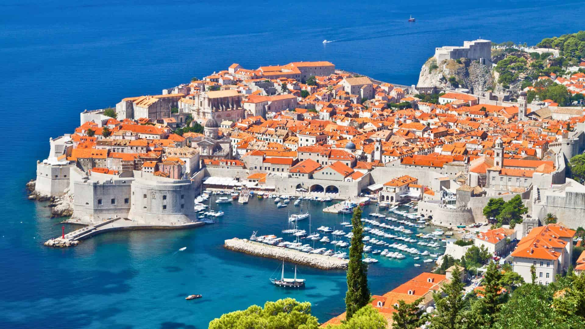 Aerial view of Dubrovnik’s old town, with orange roofs, city walls, and boats in the blue Adriatic Sea.