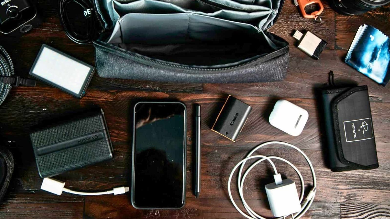 Electronics and accessories—smartphone, cables, adapters, camera battery, AirPods, zipper pouch—on a wooden surface.