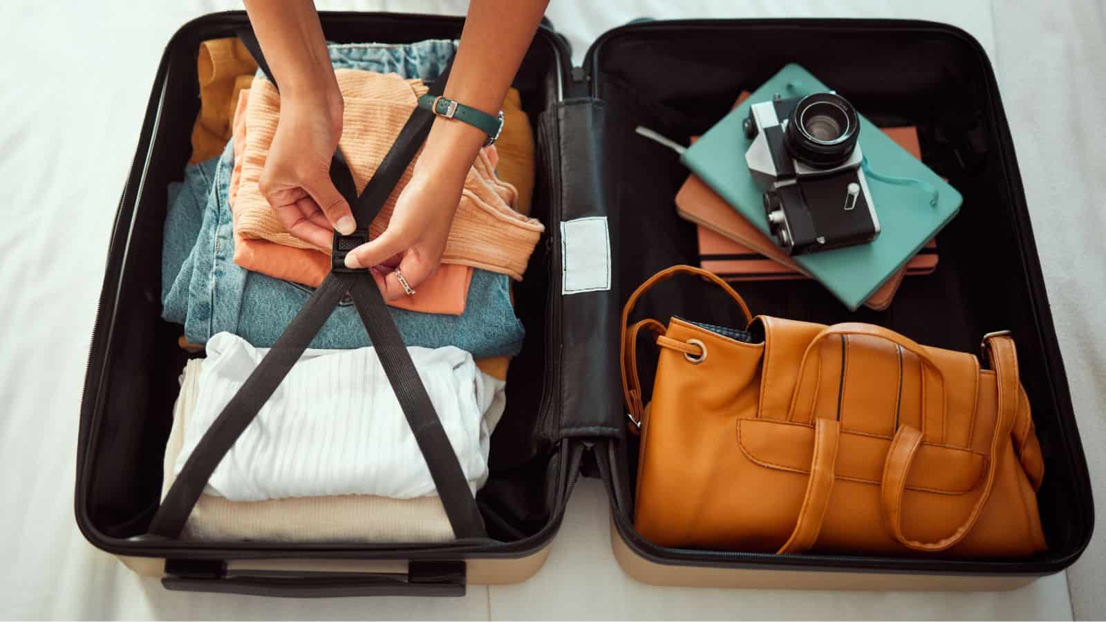 Person fastens suitcase straps over folded clothes, with a camera, notebooks, and tan handbag placed nearby.