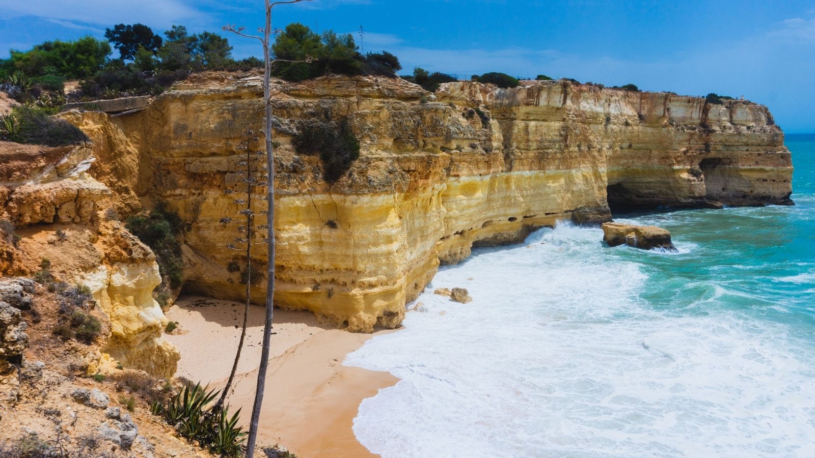 Golden limestone cliffs rising above a sandy beach and turquoise water at Praia da Marinha, Portugal.