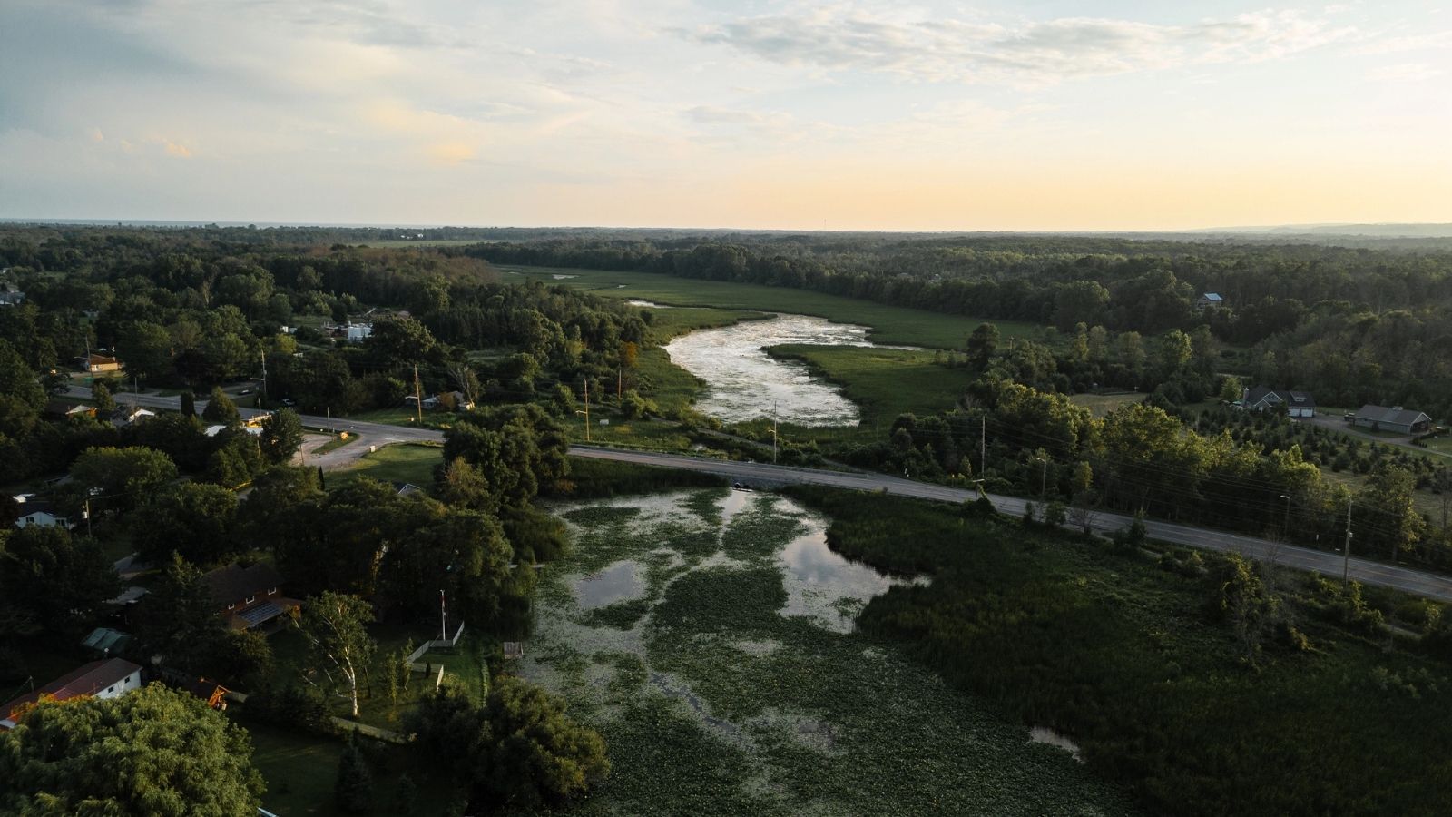 Aerial view of a rural landscape with a winding river, wetlands, dense greenery, scattered houses, and a road at sunset.