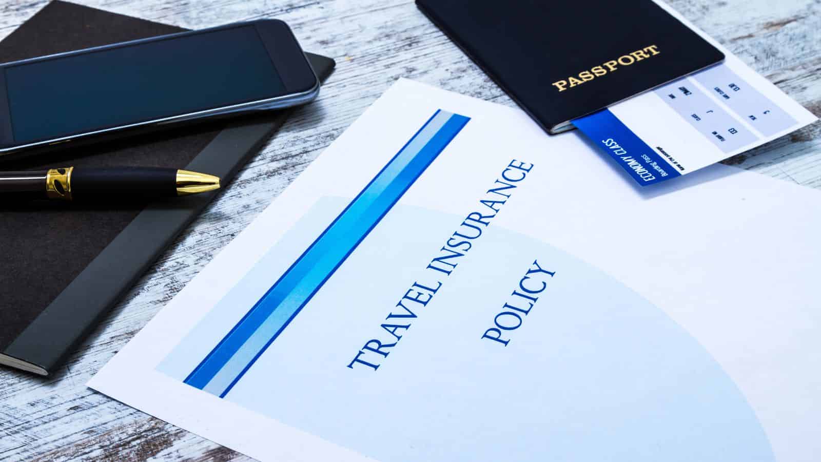 A passport, boarding pass, travel insurance document, smartphone, pen, and notebook rest on a wooden surface.