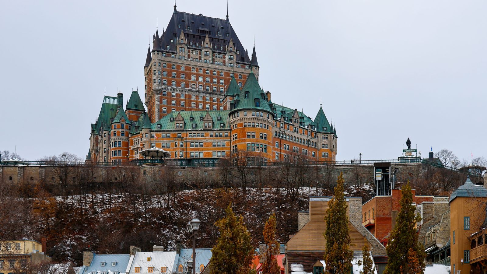 Historic chateau with green roofs and turrets overlooks a winter cityscape of trees and residential buildings.