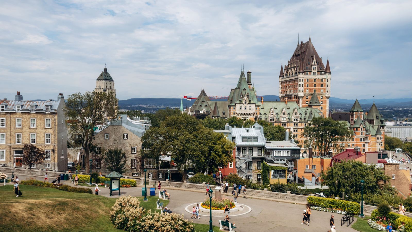 People walk in a park with historic buildings and Château Frontenac in the background under partly cloudy skies in Quebec City.