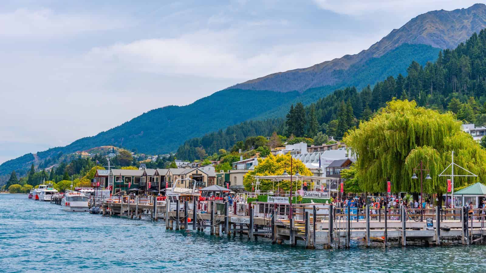 A vibrant lakeside view of Queenstown, New Zealand, with colorful buildings, a bustling dock, and green mountains in the background.