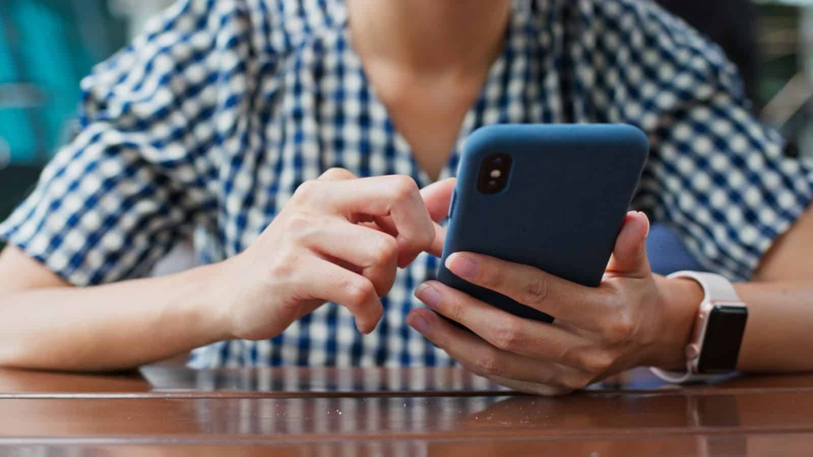 Person in a blue and white checkered shirt sits at a table, tapping on a smartphone with one hand while holding it with the other.
