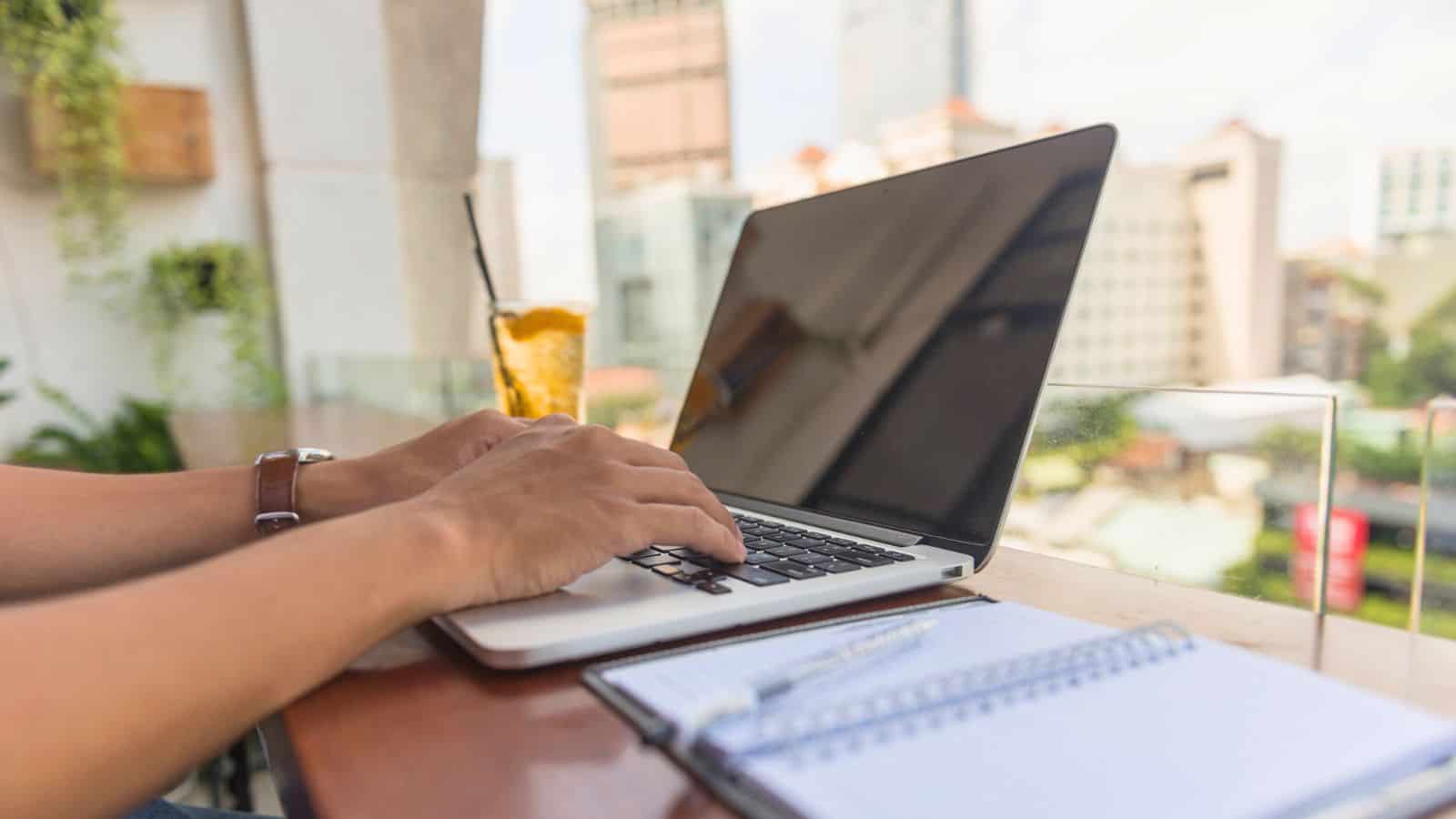 Hands type on a laptop at an outdoor table with a notebook, pen, iced tea, and city buildings in the background.