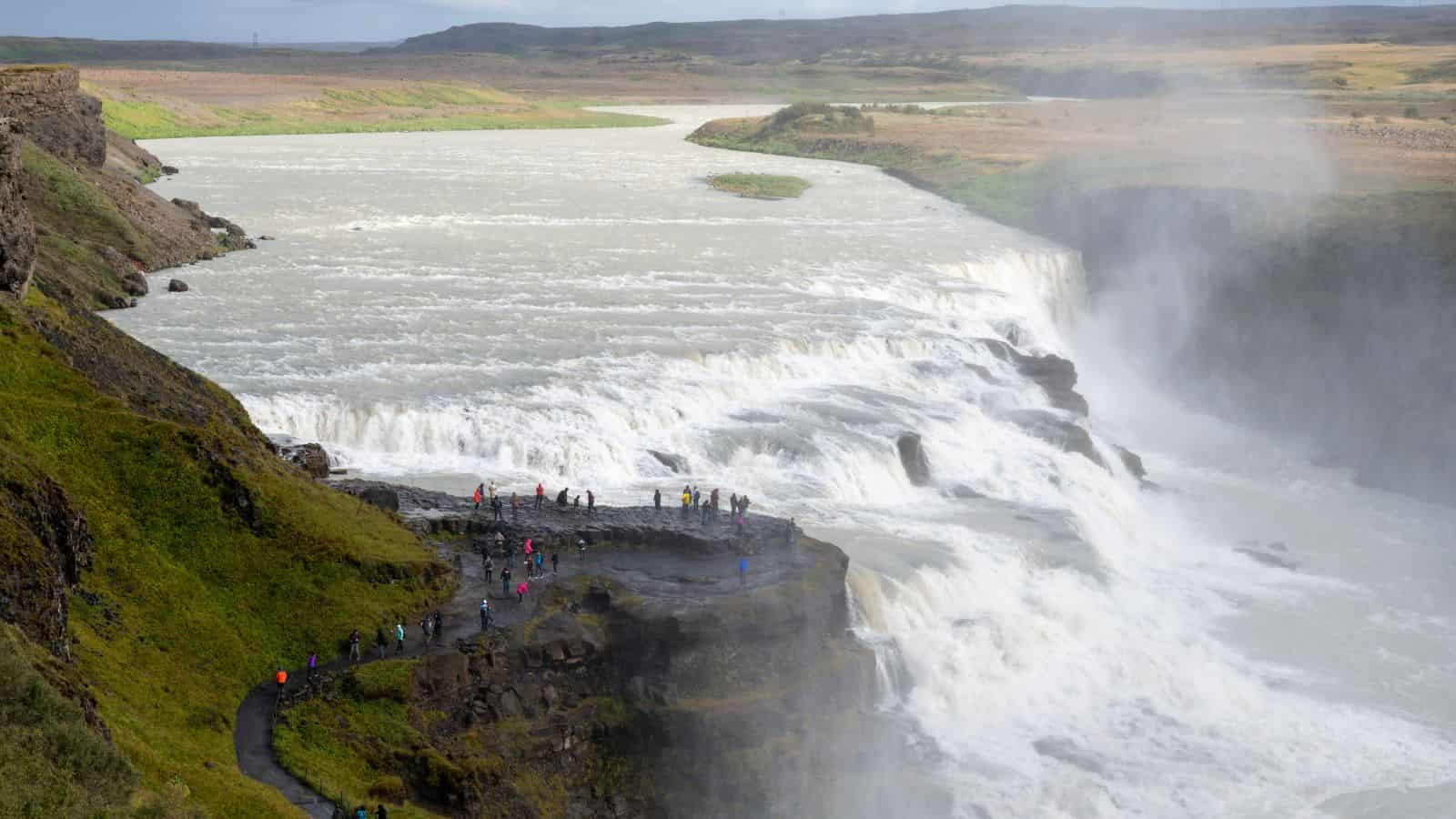 Tourists stand on a rocky path overlooking a large, multi-tiered waterfall cascading into a wide river. Mist rises from the falls, and green hills surround the area under a cloudy sky.