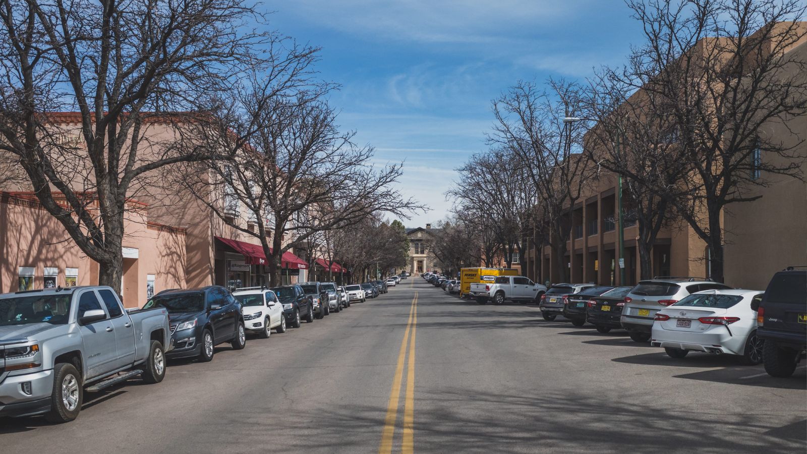 A straight two-lane street with parked cars, bare trees, and low-rise buildings under a clear blue sky.