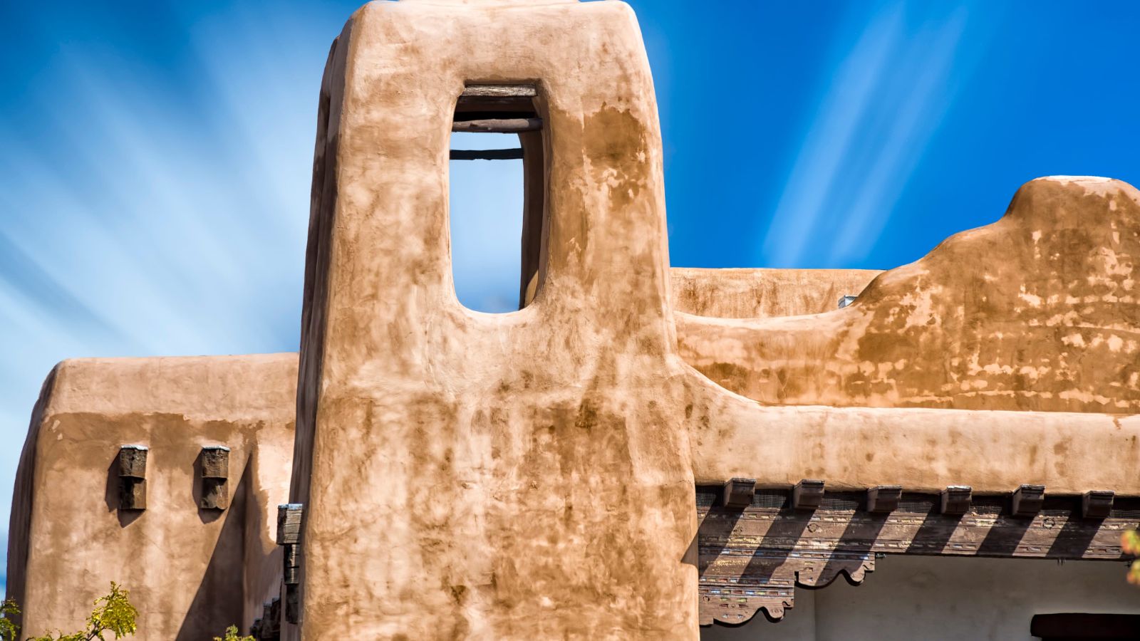 Adobe-style building with stucco walls, wooden beams, and a rectangular window below a bright blue sky.