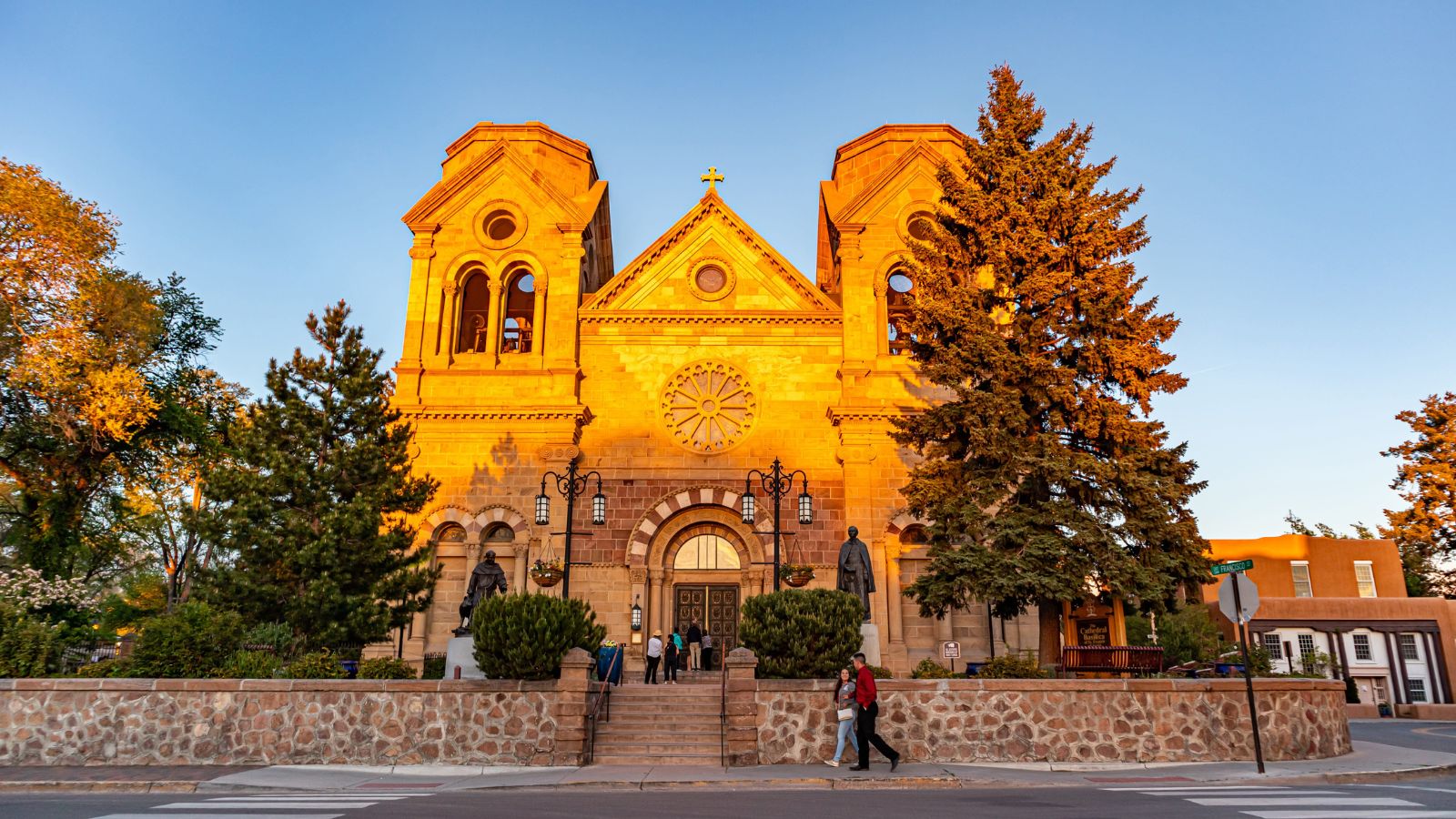 A stone cathedral with twin towers and rose window glows at sunset, as people enter, framed by trees and a stone wall.