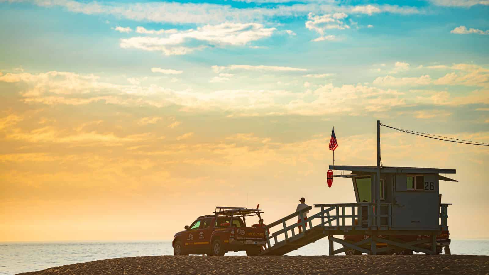 A lifeguard stands on the platform of a wooden lifeguard tower near the beach. An orange lifeguard vehicle is parked beside the tower. The sky is filled with clouds, and the ocean is visible in the background under a golden sunlight.