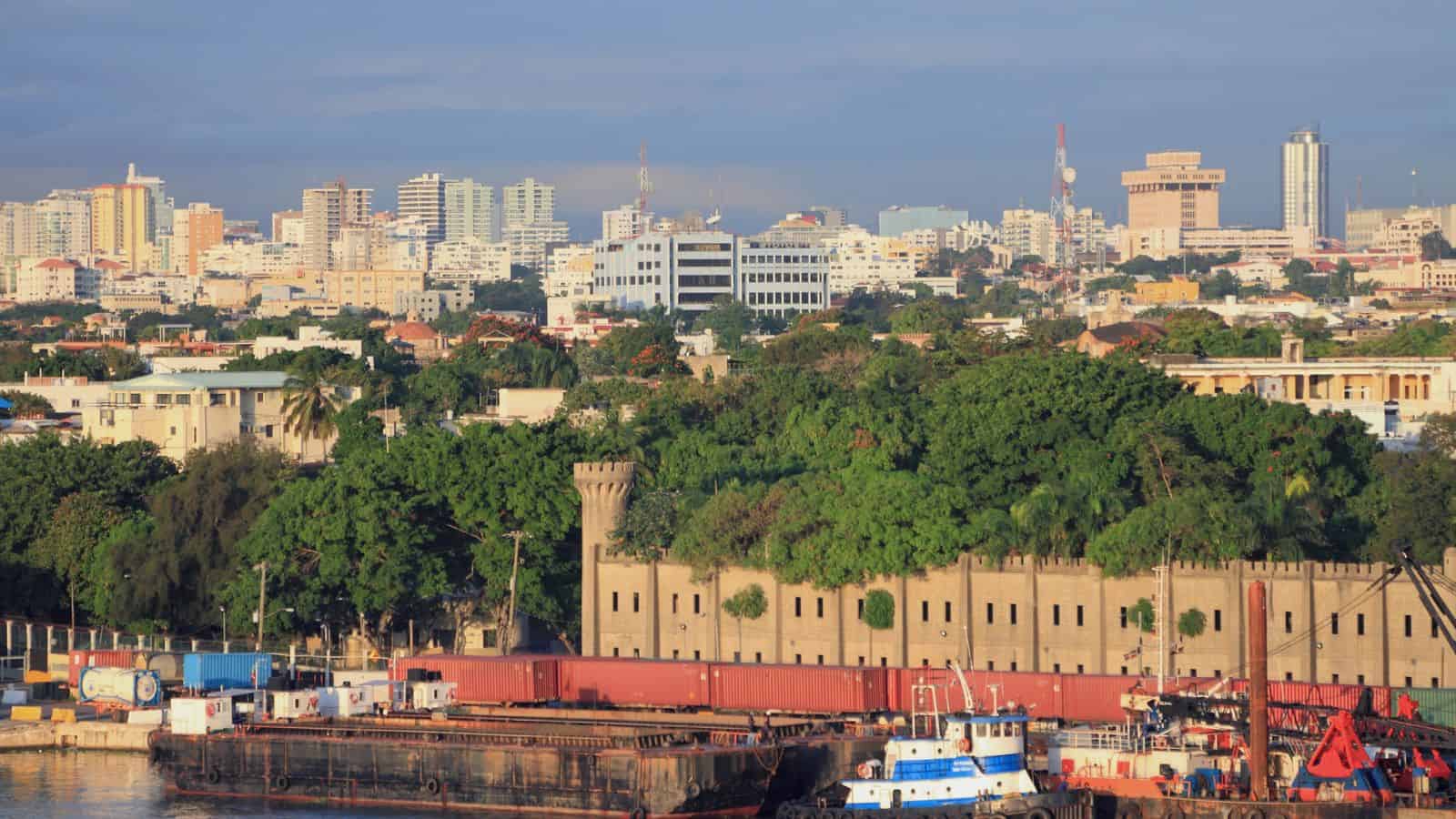 A coastal cityscape featuring a mix of modern and older buildings under a clear sky. In the foreground, there's a dock with barges and industrial equipment next to a historic fortress-like structure. Lush green trees are scattered throughout the scene.