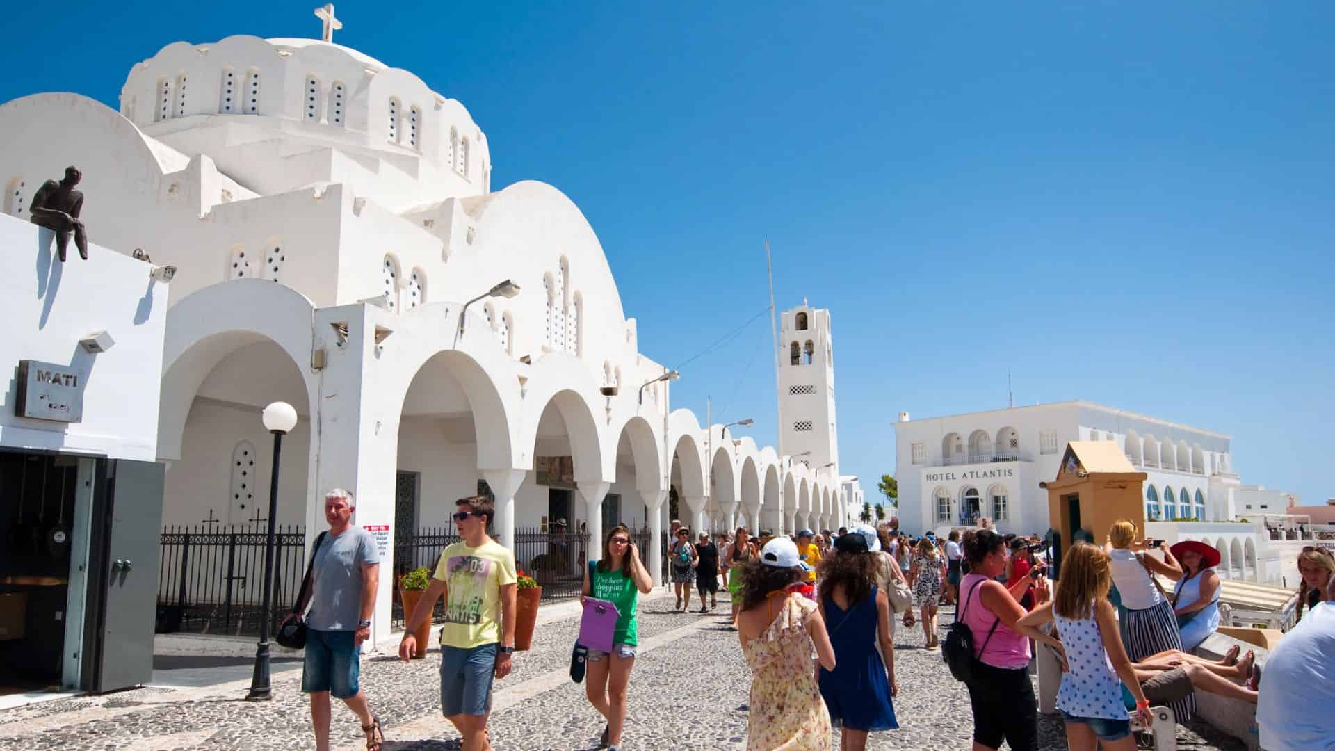 Tourists walk near a white-domed church under a clear blue sky in Santorini, Greece.