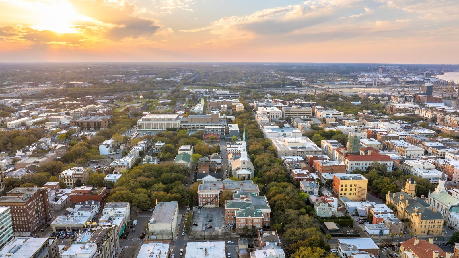Aerial sunset view of city grid with buildings, tree-lined streets, and a distant river beneath partly cloudy skies.