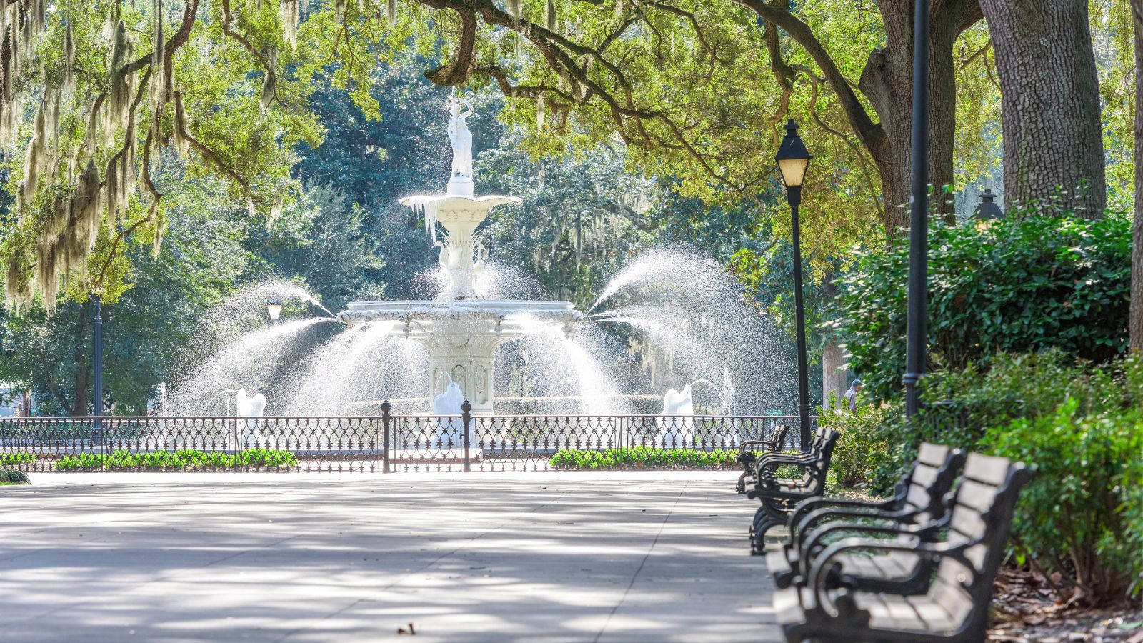 A white fountain sprays water in a tree-filled park, with benches and lamp posts along a paved walkway in front.