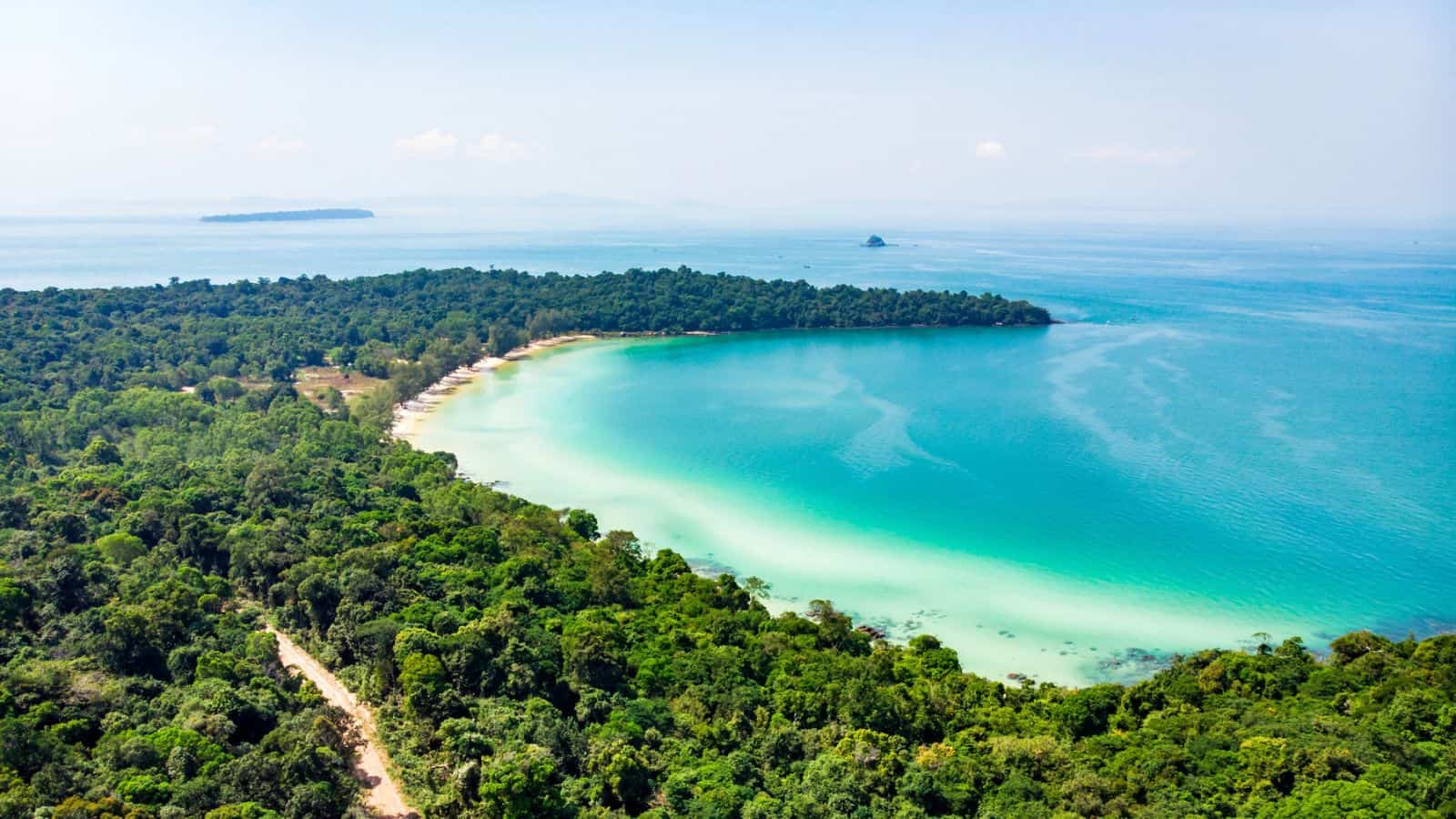 Aerial view of a curved coastline with turquoise water and white sand, bordered by dense green forest. The sea extends to the horizon under a clear sky, with small islands visible in the distance.