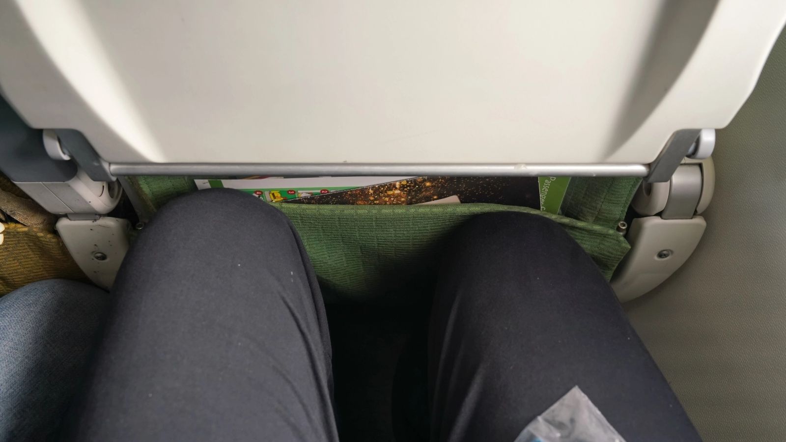 A person sits on an airplane with knees near the seat ahead and a seat pocket holding magazines visible.