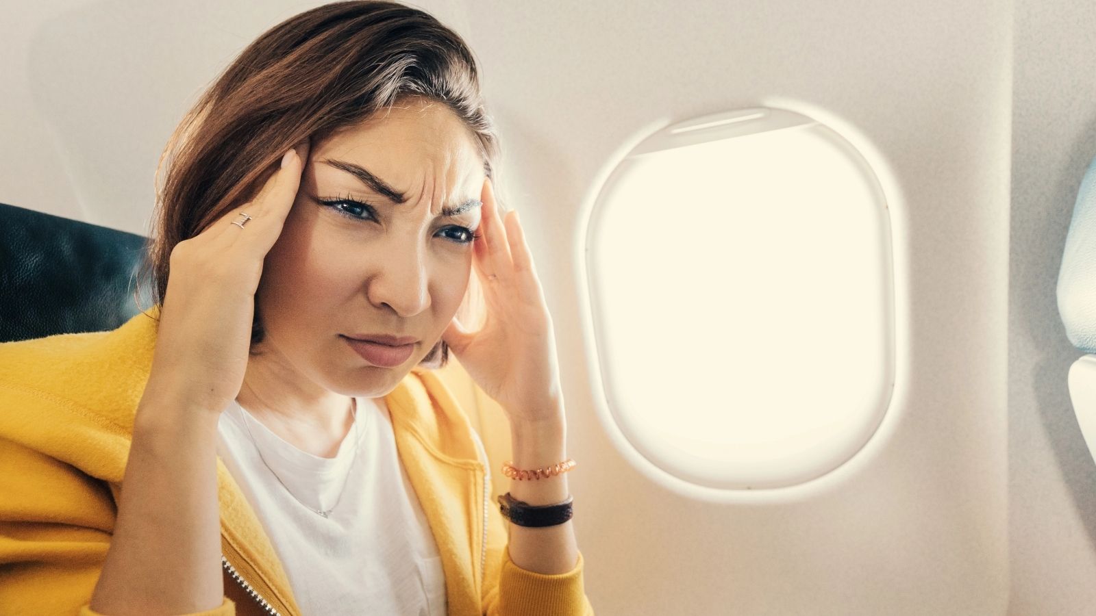 A woman by an airplane window holds her head with both hands and frowns, showing signs of discomfort or headache.