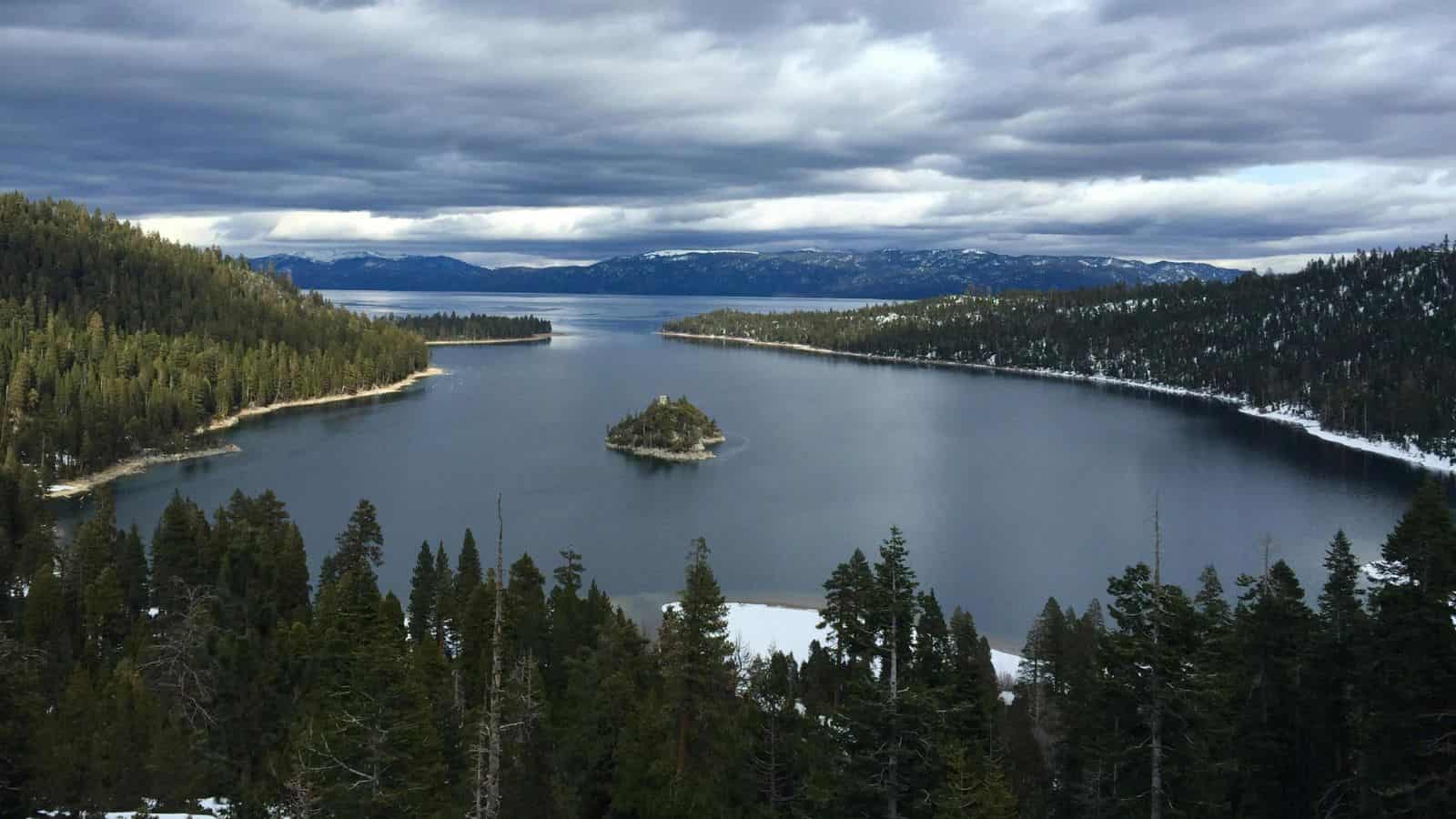 A scenic view of a lake surrounded by dense forests and mountains. An island sits in the middle of the lake. The sky is overcast with thick clouds, and patches of snow are visible on the ground and trees.