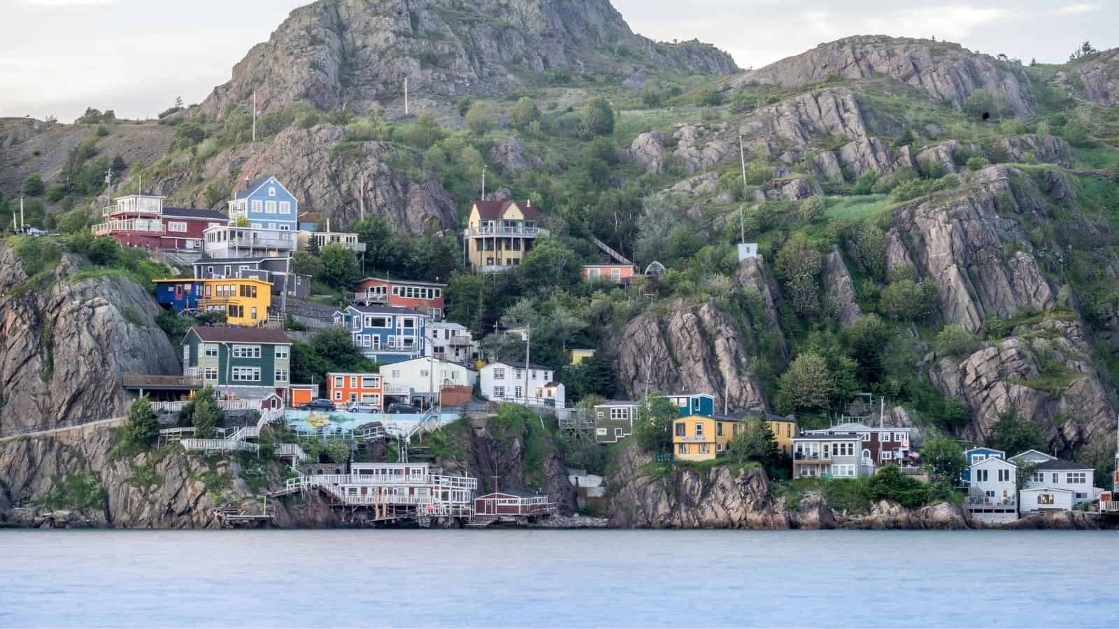 Colorful houses sit on a rocky hillside above the water, surrounded by greenery and steep cliffs.