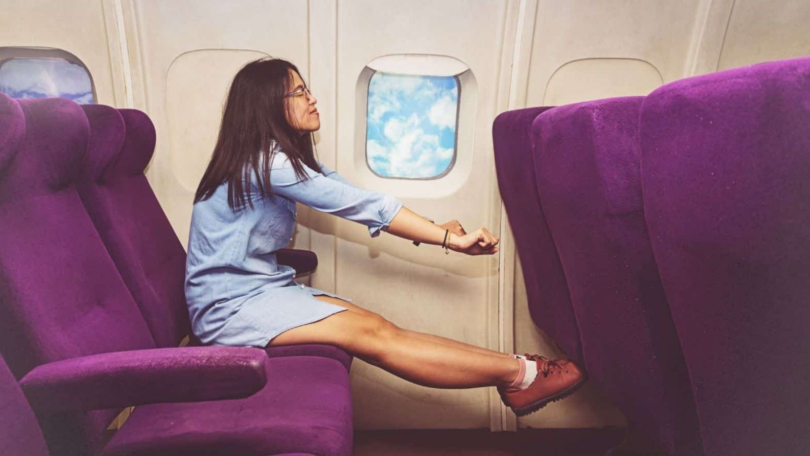 A woman in a denim dress stretches her arms and legs across purple plane seats, holding onto the seat in front of her.