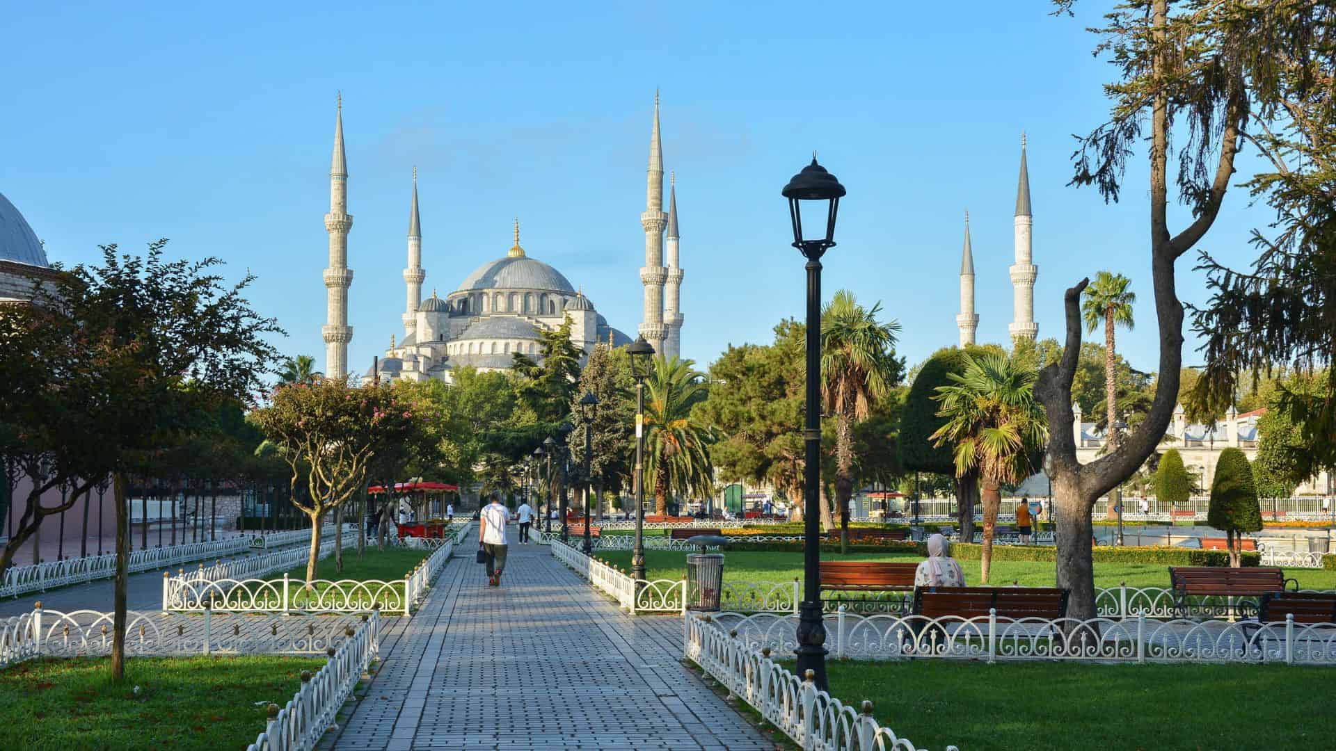 People walk along a park path lined with trees, benches, and a streetlamp toward Istanbul's Blue Mosque under a clear sky.