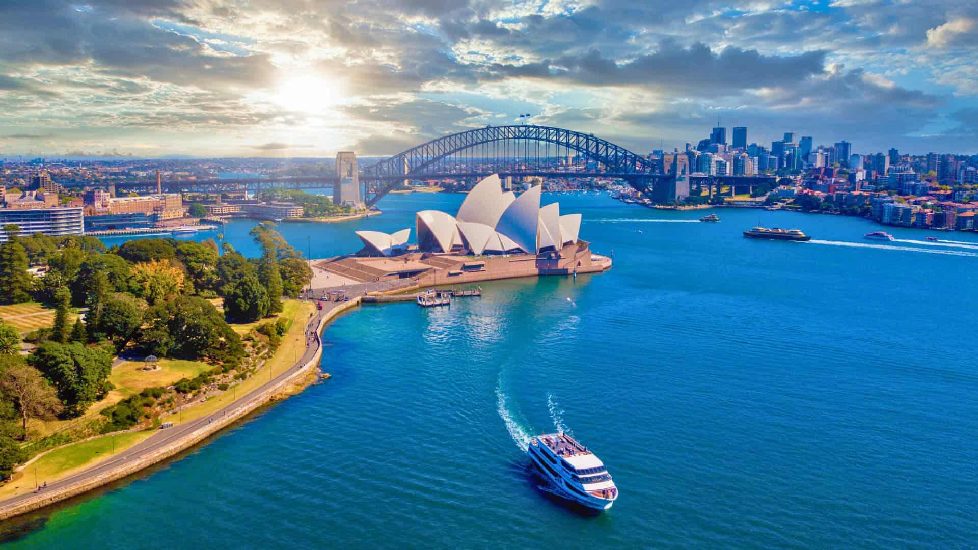 Aerial view of Sydney Harbour with the Opera House, Harbour Bridge, skyline, and boats on blue water beneath a partly cloudy sky.