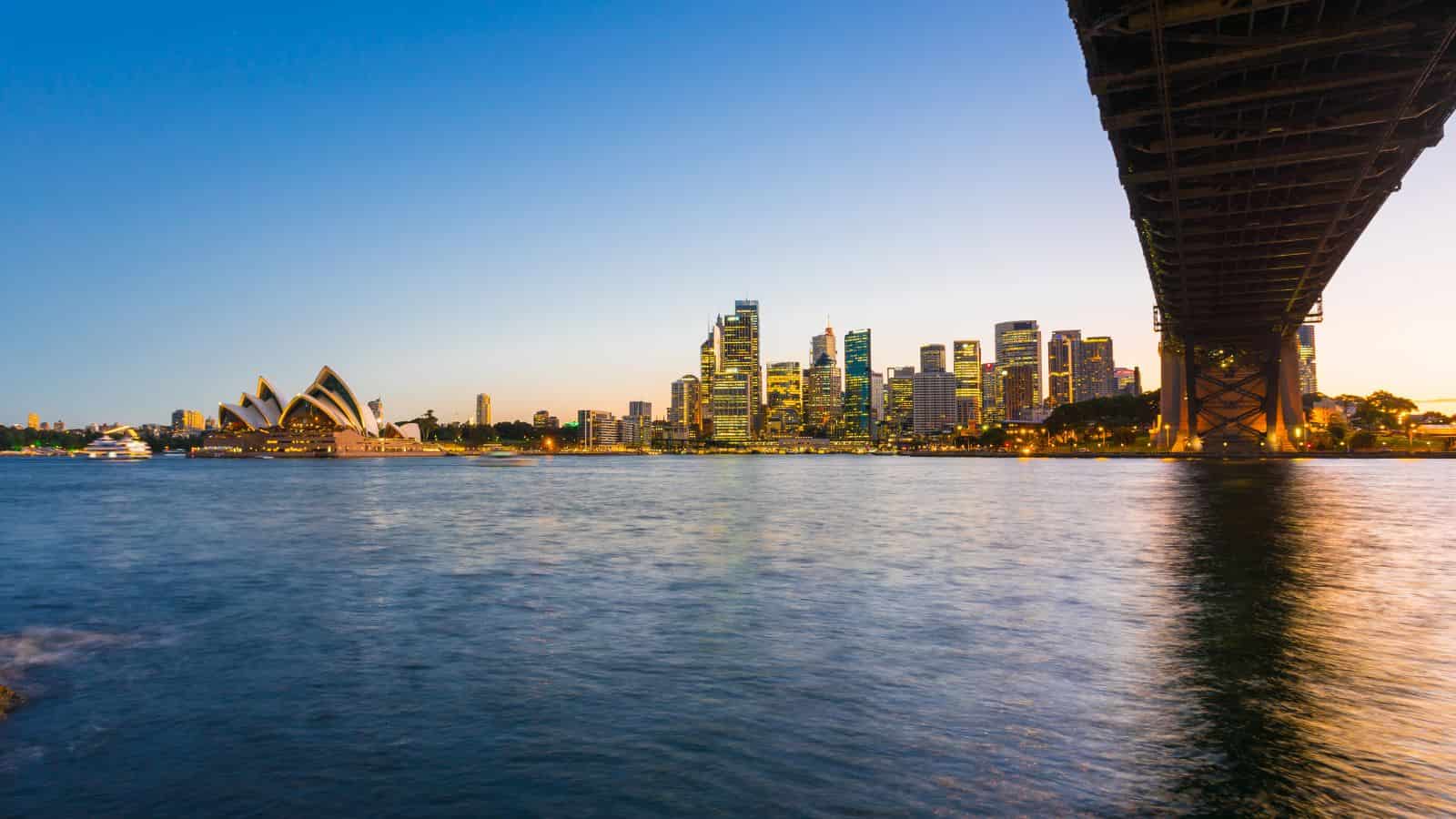 Sydney skyline at dusk with the Opera House, city buildings, and part of the Harbour Bridge visible over the water.