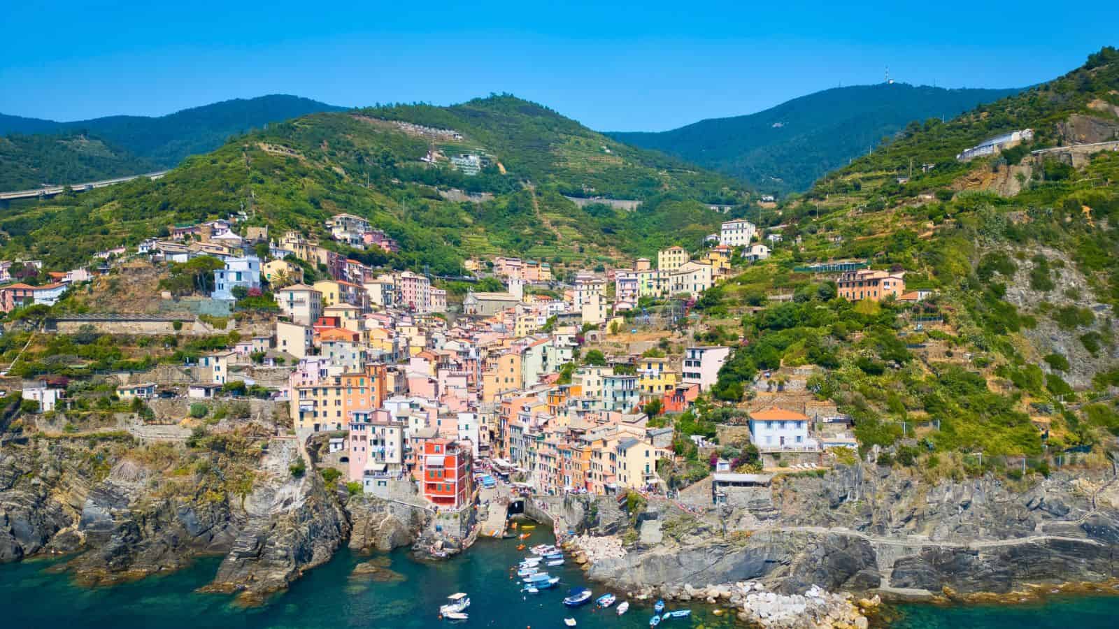 Aerial view of a coastal village with colorful buildings clustered on a hillside, surrounded by green mountains, rocky cliffs, and a small harbor with boats docked in clear blue water.