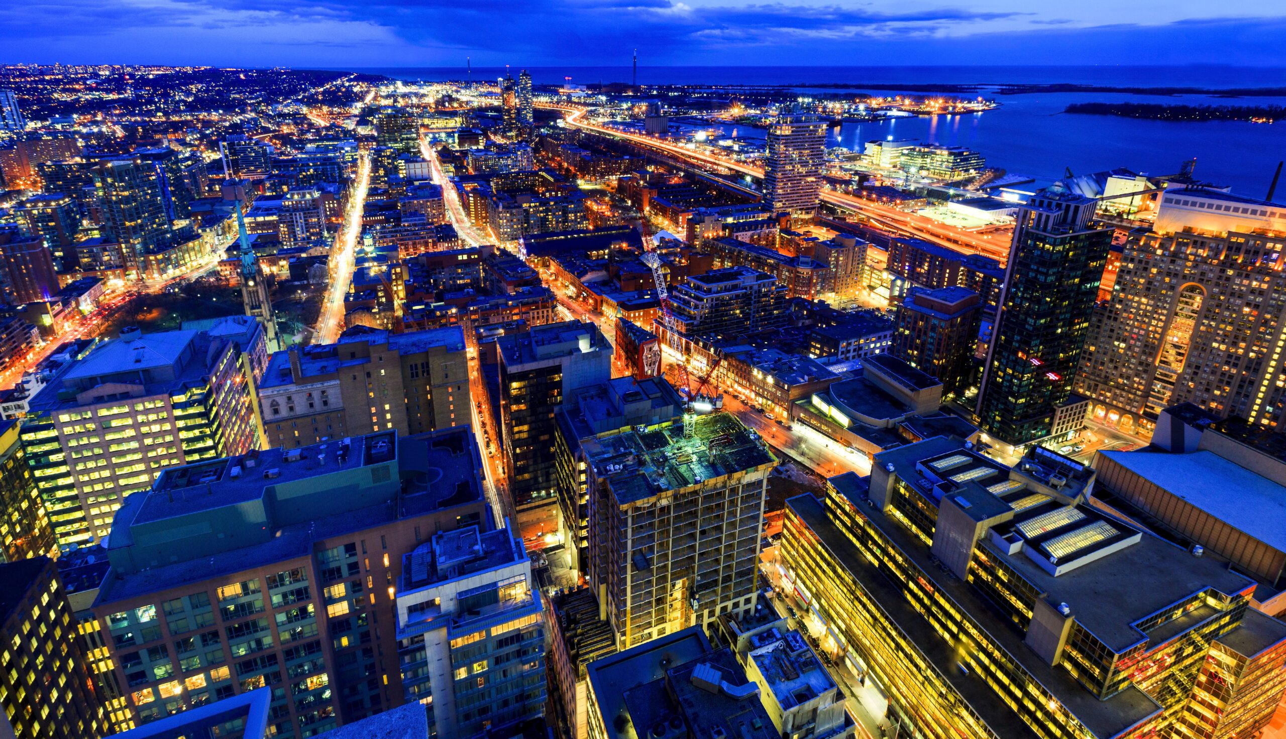 Aerial view of a city at dusk with lit streets, tall buildings, and a waterfront in the background.