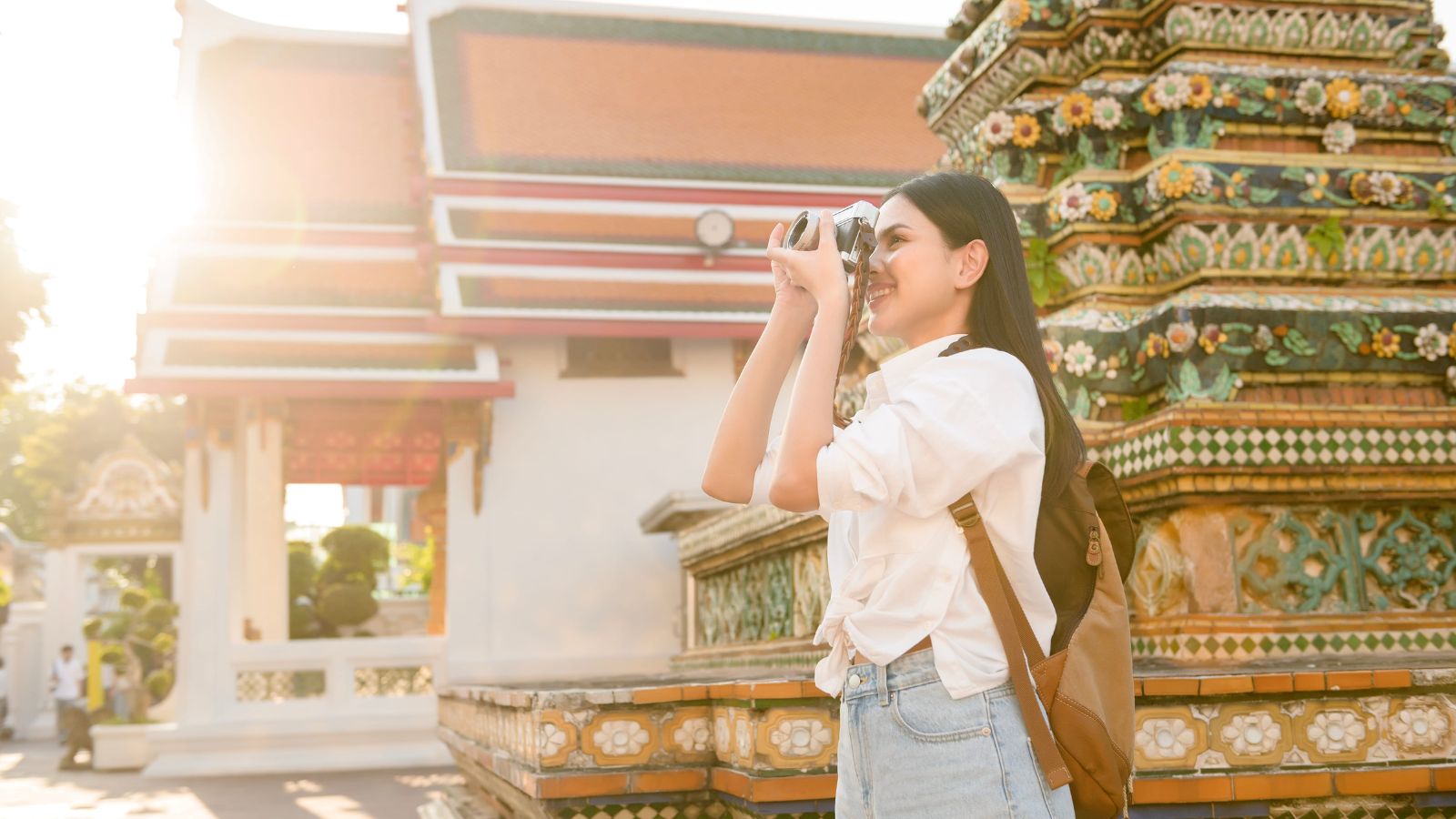 A woman wearing a backpack photographs a colorful tiled temple in bright sunlight.