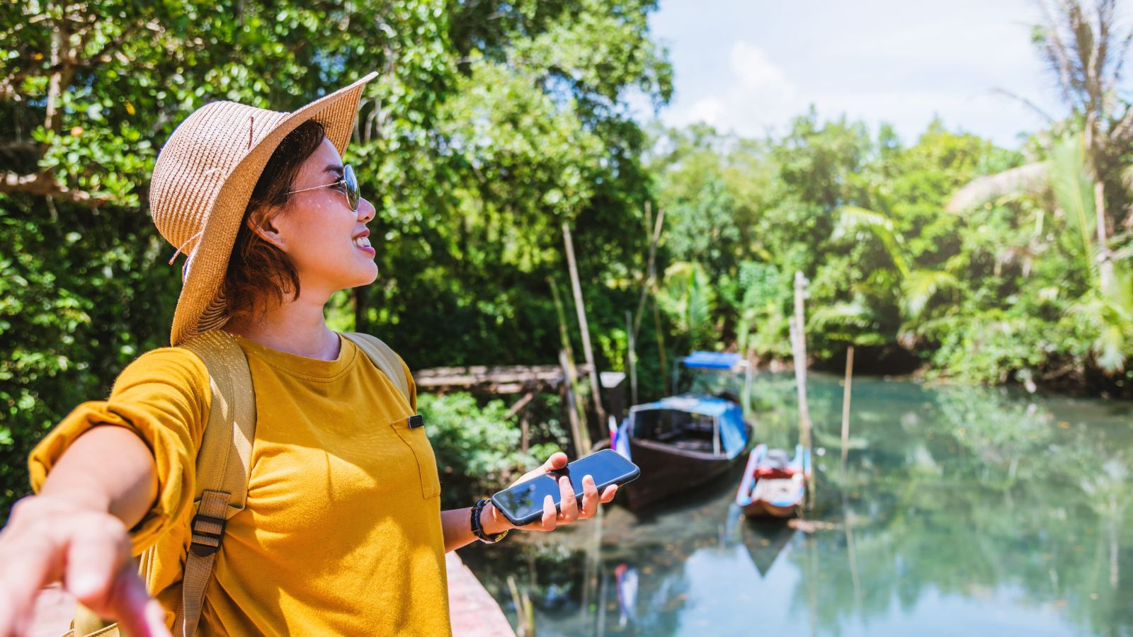 A woman in a straw hat and sunglasses points with her phone by a riverside with boats and green trees in the background.