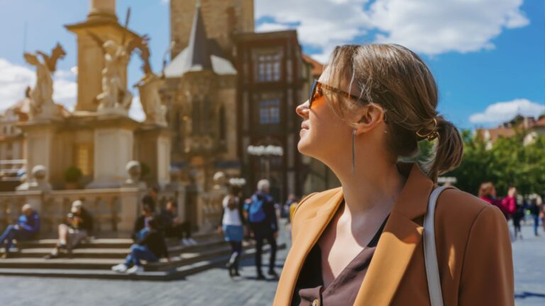 A woman in sunglasses and a brown jacket stands in a sunny city square facing historic buildings, with people in the background.