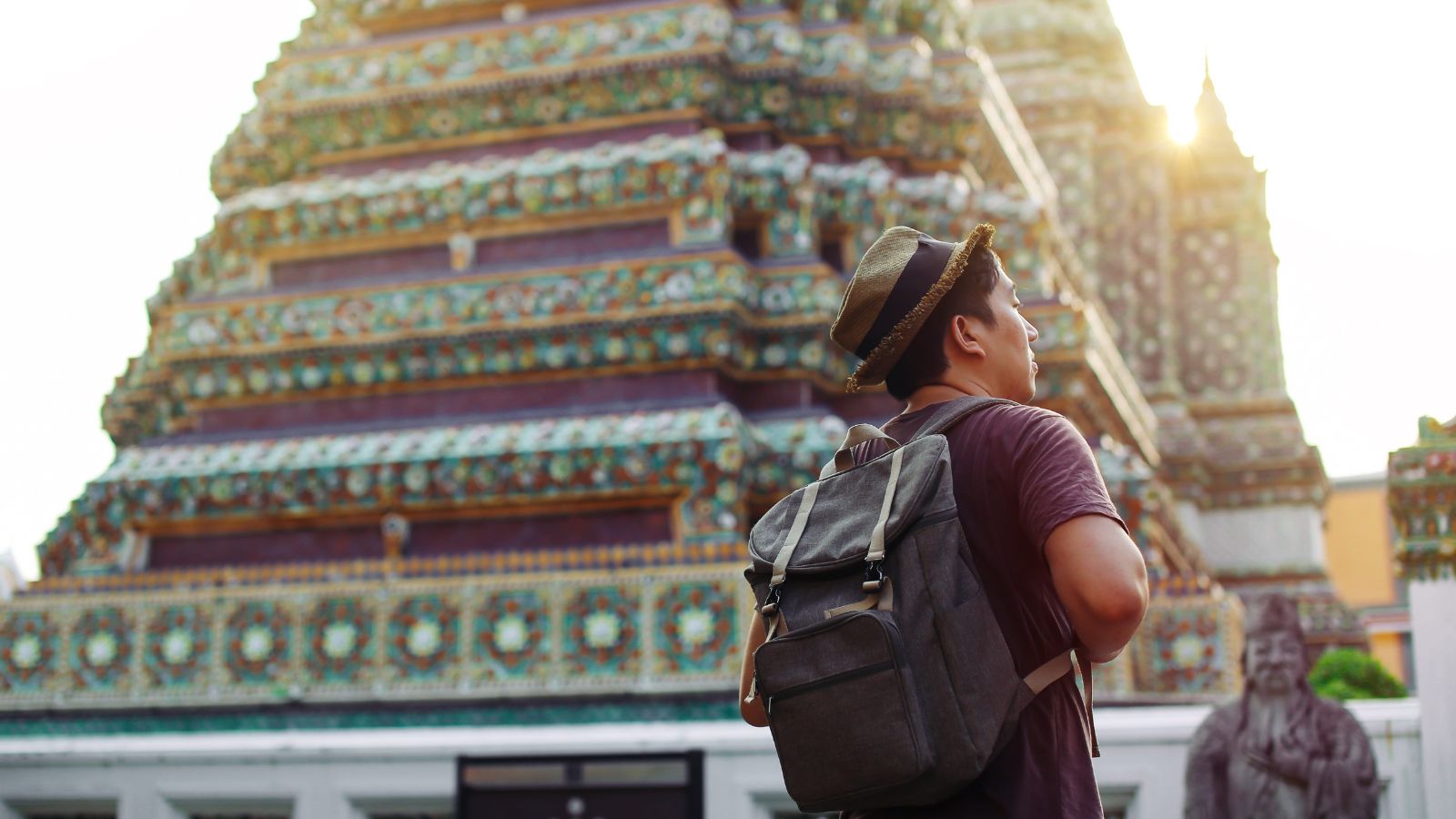 A person in a hat and backpack stands before a colorful, ornate temple on a sunny day.