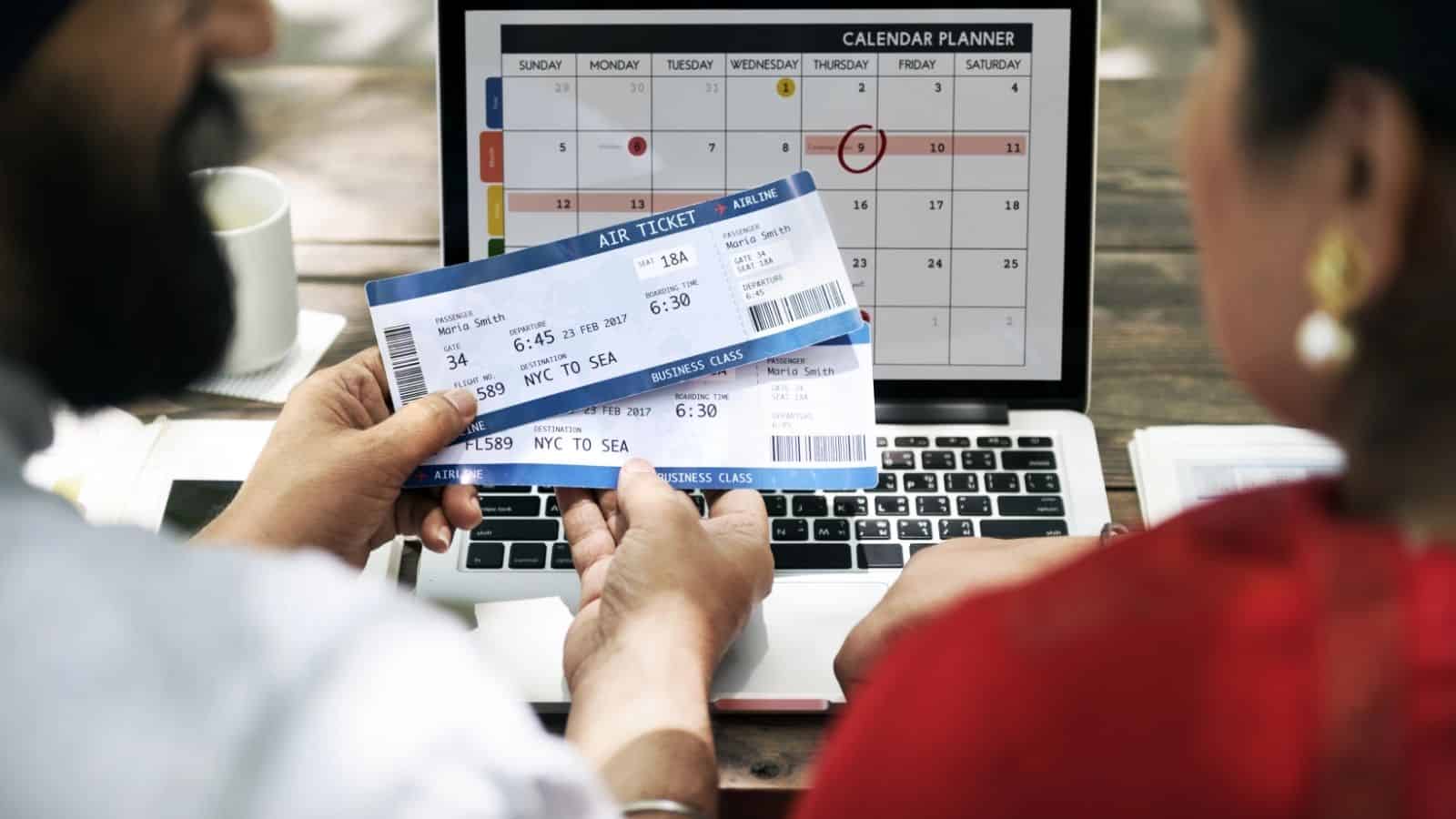 Two people hold airline tickets by a laptop showing a calendar planner, suggesting travel planning.